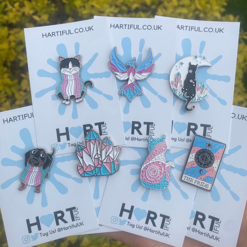 May include: A collection of enamel pins on backing cards. The pins feature various designs, including a cat, a phoenix, a dachshund, a crystal, a cat on a crescent moon, and a tarot card. The pins have a color scheme of pink, blue, and white. The cards have the text "HARTIFUL.CO.UK".