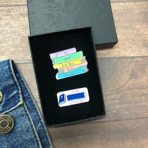 Pode incluir: Dois pinos de esmalte em uma caixa preta. Um pino apresenta uma pilha de livros coloridos com o texto "Sim, preciso de todos esses livros (e mais)". O outro pino é azul e branco e diz "Alimentado por livros".