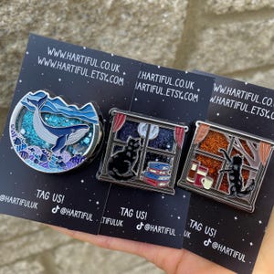 Puede incluir: Tres pines de esmalte sobre tarjetas negras. Un pin presenta una ballena en una escena marina, otro un gato negro junto a una ventana con libros, y el tercero un gato junto a una ventana con una taza. Las tarjetas tienen el texto "TAG US!" y direcciones web.