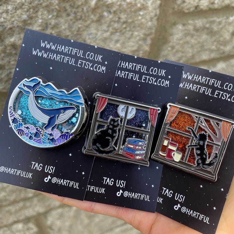 Puede incluir: Tres pines de esmalte sobre tarjetas negras. Un pin presenta una ballena en una escena marina, otro un gato negro junto a una ventana con libros, y el tercero un gato junto a una ventana con una taza. Las tarjetas tienen el texto "TAG US!" y direcciones web.