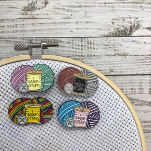以下が含まれることがあります： 糸巻きと「I Make Stuff」という文字とボタンが描かれたエナメルピンが4つ。それぞれのピンは、ピンクとグリーン、レッドとグレー、レインボー、ブルーとパープルの異なる色の背景になっています。