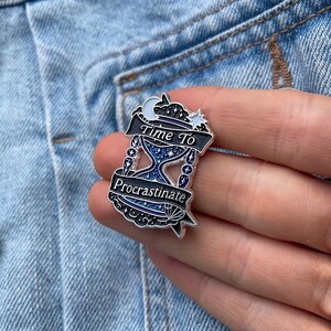 May include: A silver-colored enamel pin with the text "Time To Procrastinate". The pin features an hourglass design with blue and white details, a crescent moon, and a star. The pin is set against a denim background.