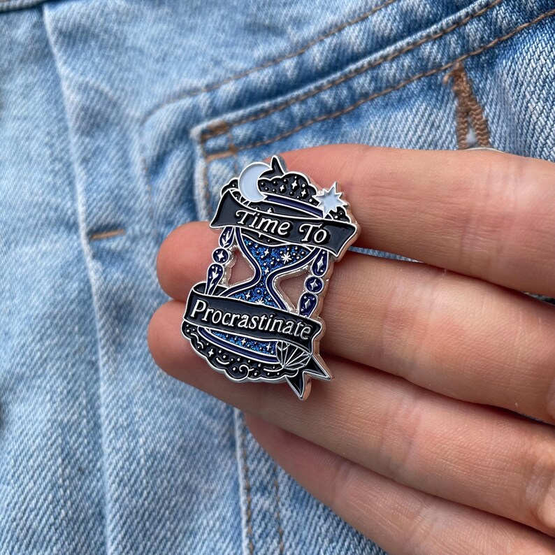 May include: A silver-colored enamel pin with the text "Time To Procrastinate". The pin features an hourglass design with blue and white details, a crescent moon, and a star. The pin is set against a denim background.