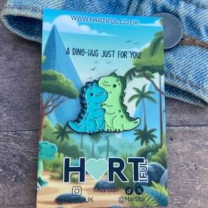 May include: A green and blue enamel pin featuring two cartoon dinosaurs hugging. The pin is on a card with the text "A Dino-Hug Just For You!" and the logo "HORT" with a heart in the middle.