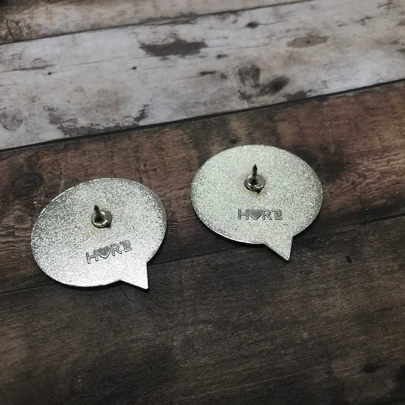 May include: Two silver metal pins shaped like speech bubbles. Each pin has the word "Hore" written on it with a small heart symbol in between the letters.
