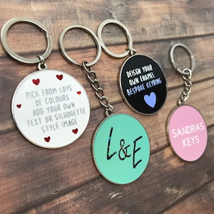 May include: Four different enamel keychains with different designs. One keychain is white with red hearts and says "Pick from lots of colours add your own text or silhouette style image". Another keychain is black with a light blue heart and says "Design your own enamel bespoke keyring". The third keychain is light green with black glitter and says "L&E". The fourth keychain is pink and says "Sandras Keys".