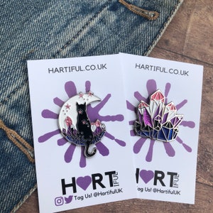 May include: Two enamel pins on white cards with purple splatter paint. The first pin features a black cat sitting on a crescent moon with pink and purple crystals. The second pin features a cluster of pink, purple, and blue crystals. Both cards have the text "HARTIFUL.CO.UK" and "HEARTIFUL" with a heart symbol.