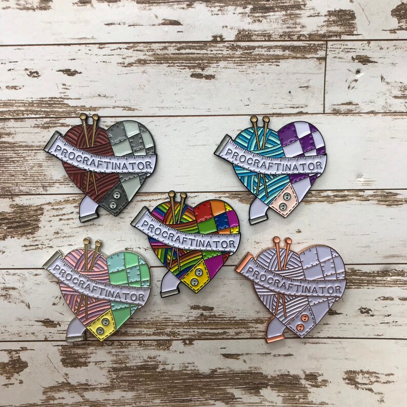 May include: Five enamel pins shaped like hearts with a yarn ball and knitting needles inside. Each pin has a white banner with the text "PROCRASTINATOR" on it. The hearts are different colors and patterns: red and white checkered, purple and blue striped, rainbow, green and pink checkered, and white with a light blue and pink pattern.