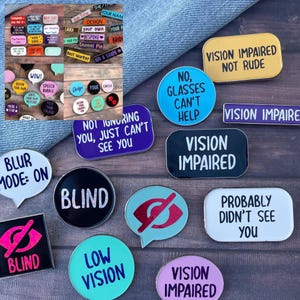 May include: A selection of enamel pins with diverse designs and text. The pins display phrases such as "Vision Impaired," "Blind," and "Blur Mode: On." They come in various shapes and colours, including black, white, blue, and yellow.