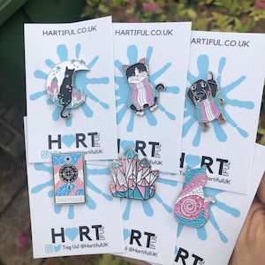 May include: A collection of enamel pins featuring cat and dog designs, crystals, and a tarot card. The pins are in various colors, including black, pink, and blue, and are displayed on white cards with the text "HARTIFUL.CO.UK".