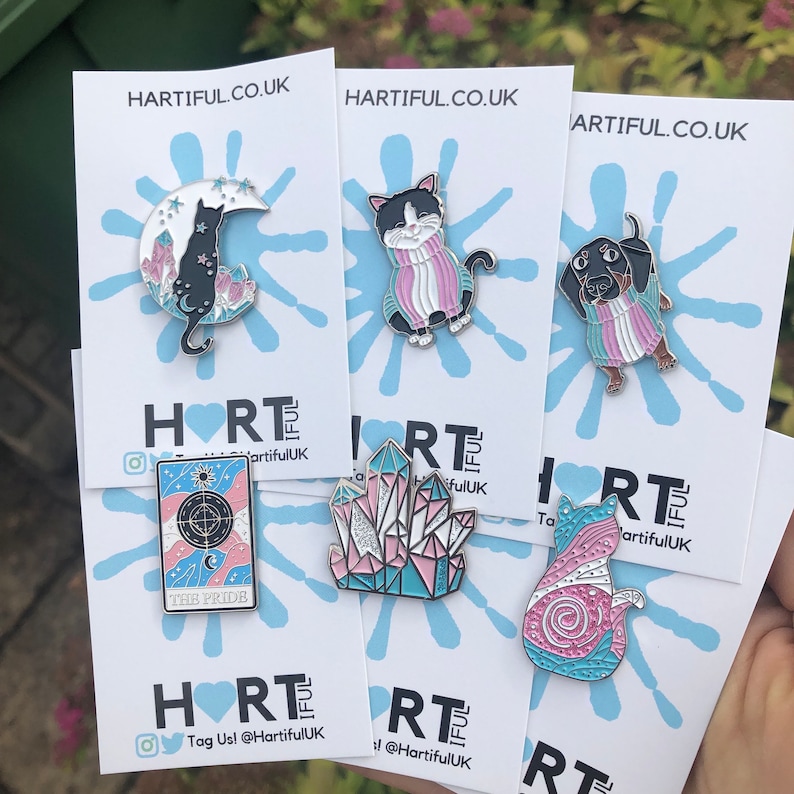 May include: A collection of enamel pins featuring cat and dog designs, crystals, and a tarot card. The pins are in various colors, including black, pink, and blue, and are displayed on white cards with the text "HARTIFUL.CO.UK".