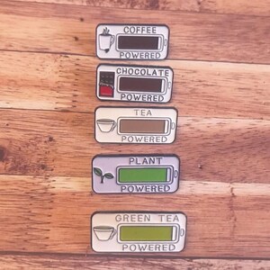 K&ouml;nnte beinhalten: F&uuml;nf Emaille-Pins mit einem Batterie-Symbol und dem Text "COFFEE POWERED", "CHOCOLATE POWERED", "TEA POWERED", "PLANT POWERED" und "GREEN TEA POWERED".