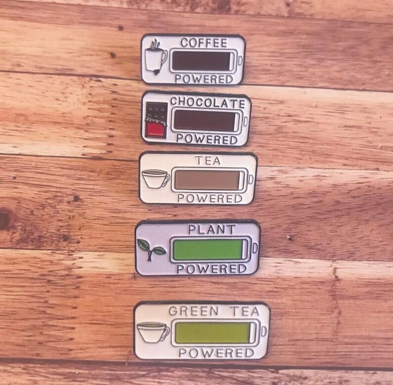K&ouml;nnte beinhalten: F&uuml;nf Emaille-Pins mit einem Batterie-Symbol und dem Text "COFFEE POWERED", "CHOCOLATE POWERED", "TEA POWERED", "PLANT POWERED" und "GREEN TEA POWERED".
