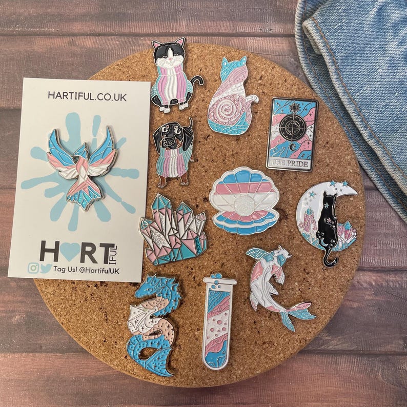 May include: A collection of enamel pins on a corkboard. The pins feature various designs, including a phoenix, cats, a dog, crystals, a seashell, a cat on a crescent moon, a koi fish, a dragon, and a test tube. The pins incorporate blue, pink, and white colors.