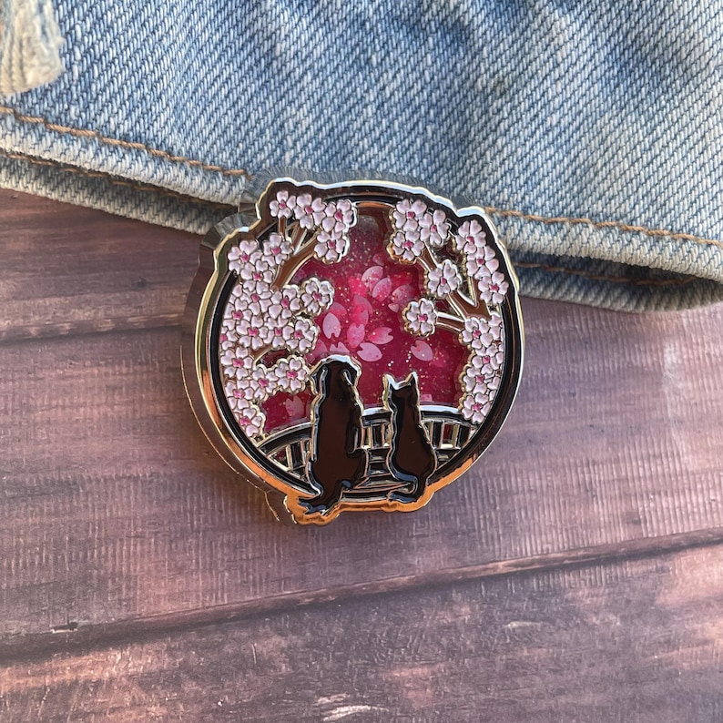 Puede incluir: Un pin redondo de esmalte dorado que presenta la silueta de un perro y un gato bajo un cerezo en flor. El fondo es de un rosa vibrante con p&eacute;talos cayendo. El pin est&aacute; sobre un fondo de mezclilla.
