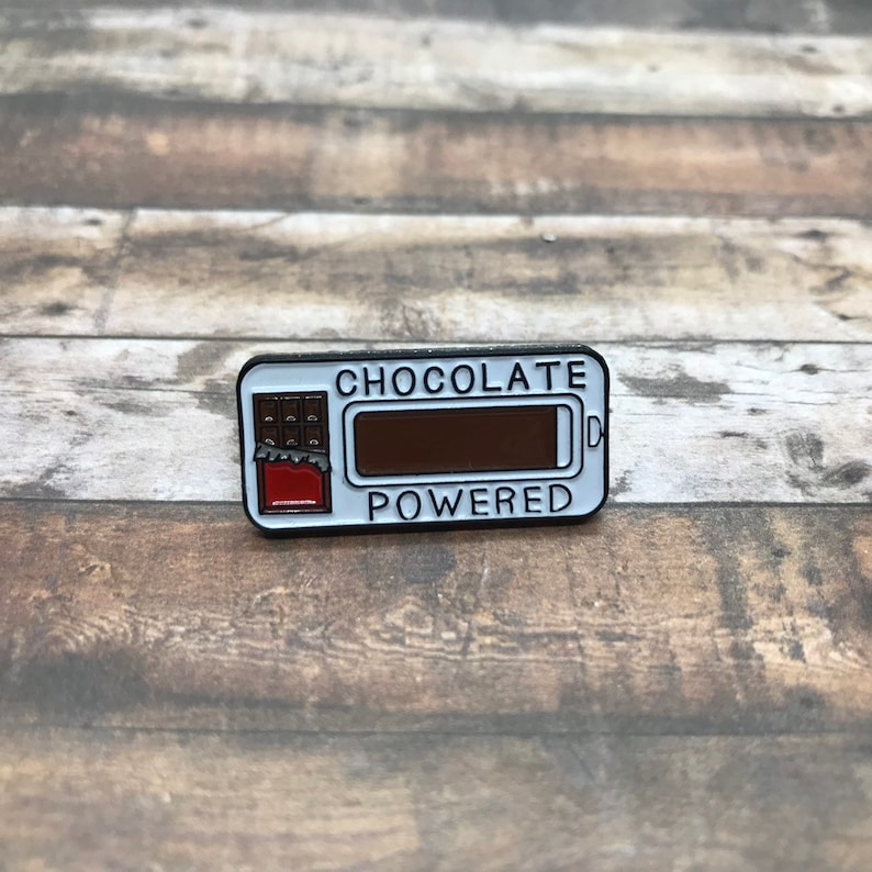K&ouml;nnte beinhalten: Eine wei&szlig;e Emaille-Anstecknadel mit einer Schokoladentafel-Grafik und dem Text "CHOCOLATE POWERED".
