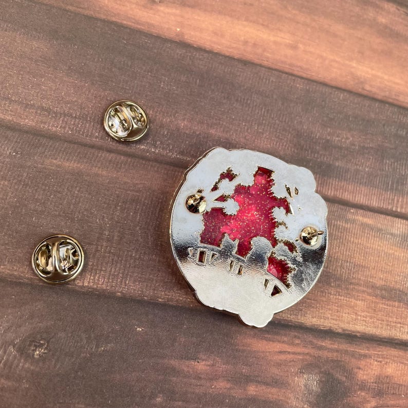 Puede incluir: Un pin de color plateado con un centro de purpurina roja y un dise&ntilde;o decorativo recortado. Se ven dos cierres de pin dorados, junto con la parte posterior del pin. El pin est&aacute; sobre una superficie de madera.