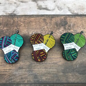 May include: Three enamel pins featuring yarn balls and knitting needles. Each pin has a white banner that reads "Just One More Row."