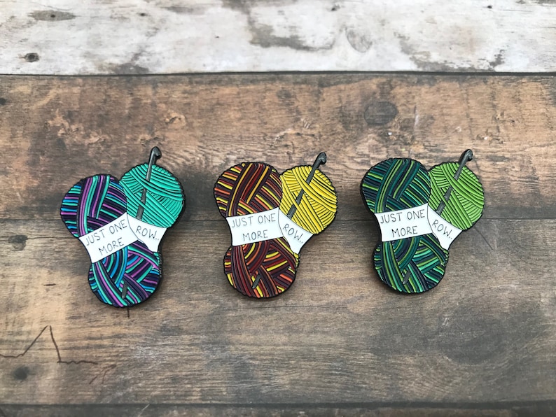 May include: Three enamel pins featuring yarn balls and knitting needles. Each pin has a white banner that reads "Just One More Row."