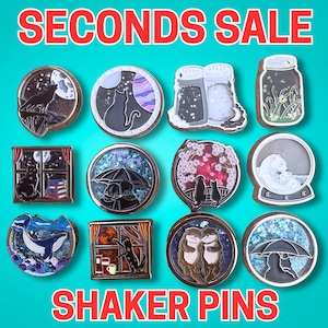 May include: A collection of twelve decorative shaker pins featuring various designs including a wolf, cats, salt and pepper shakers, a jar, a whale, otters, and figures under umbrellas, with the text SECONDS SALE and SHAKER PINS displayed.