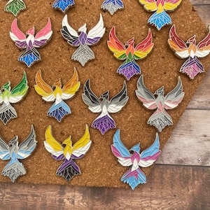 May include: A collection of phoenix-shaped enamel pins in various colors, including rainbow, pink, blue, and black. Each pin features a detailed design with a silver outline and glitter accents. The pins are displayed on a corkboard.