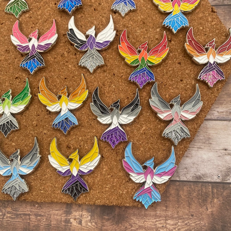May include: A collection of phoenix-shaped enamel pins in various colors, including rainbow, pink, blue, and black. Each pin features a detailed design with a silver outline and glitter accents. The pins are displayed on a corkboard.