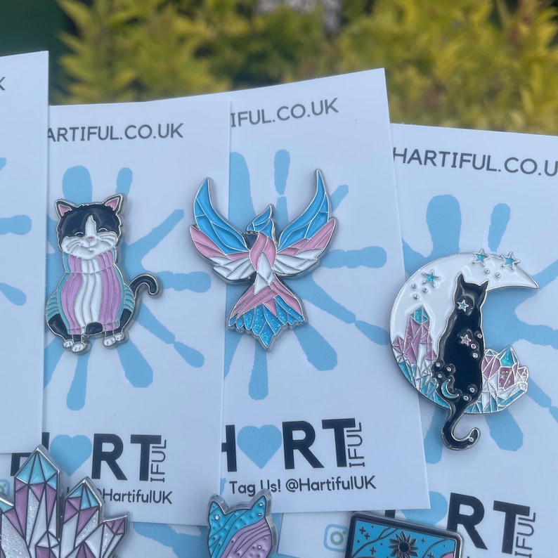 May include: A collection of enamel pins featuring various designs. One pin depicts a black and white cat wearing a pink and blue scarf. Another shows a bird with blue, pink, and white wings. The third pin features a black cat on a crescent moon with crystals.