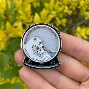 May include: A silver-colored snow globe pin featuring a mother polar bear and her cub. The bears are white with black outlines, set against a snowy background. The base of the pin is black with blue accents.