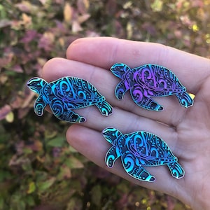 May include: Three iridescent enamel pins of sea turtles with intricate designs. The turtles are in shades of blue, purple, and green.