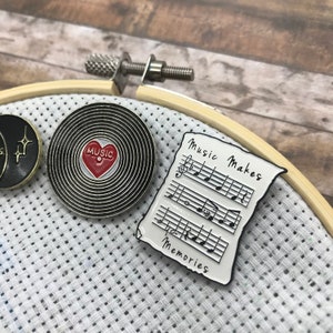 May include: Two enamel pins, one with a black and white record design with a red heart that says "MUSIC" and the other with a white sheet of music with the text "Music Makes Memories".