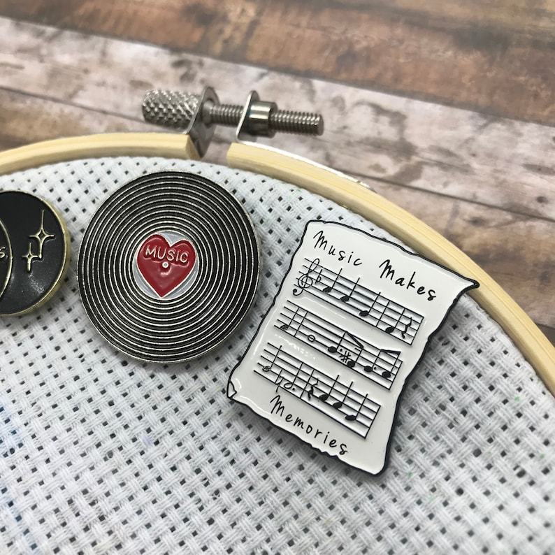 May include: Two enamel pins, one with a black and white record design with a red heart that says "MUSIC" and the other with a white sheet of music with the text "Music Makes Memories".