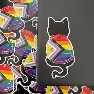 May include: Black cat sticker with a white outline, wearing a sweater with a rainbow Pride flag design. The sticker has a circular yellow accent. Additional stickers with the same design are visible.