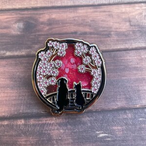 Puede incluir: Un pin redondo de esmalte dorado que presenta una silueta negra de un perro y un gato sentados juntos, enmarcado por ramas de cerezos en flor. El fondo es rosa vibrante con detalles de purpurina, sobre una superficie de madera oscura.