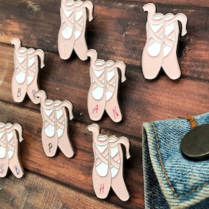 Peut inclure: Ensemble de dix épingles en émail rose en forme de ballerine avec des lettres différentes sur chaque chaussure. Les lettres épellent le mot "ballerina".
