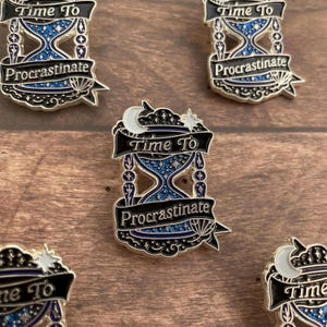 May include: Enamel pins with a black and silver border, featuring an hourglass design with blue glitter and the words "Time To Procrastinate." The pins have a crescent moon and star detail at the top.
