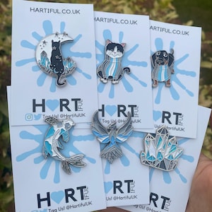 Pode incluir: Um conjunto de pinos de esmalte com um gato preto sentado em uma lua crescente com cristais, um gato preto e branco usando uma echarpe listrada azul e branca, um dachshund usando uma echarpe listrada azul e branca, um peixe koi azul e branco, um pássaro azul e branco com as asas abertas e um grupo de cristais azuis e brancos. Os pinos estão todos em cartões brancos com o texto "HARTIFUL.CO.UK" e "Tag Us! @HartifulUK" impresso neles.
