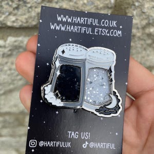 May include: A black enamel pin featuring two salt shakers, one black and one filled with glitter. The pin is on a black card with the text "TAG US!" and social media handles. The pin is held by a hand.