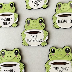 Puede incluir: Una colección de pines de esmalte de ranas de color verde claro, cada una sosteniendo una taza de café blanca. Las tazas muestran texto en negro con varios pronombres, incluyendo HE/SHE/THEY, ZE/ZIR/ZEM, THEY/THEM, ANY PRONOUNS, SHE/HER y THEY/SHE/HE.