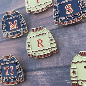 May include: Close-up of several enamel pins shaped like holiday sweaters. The pins are in shades of blue and green, with letters and star accents. The letters include M, R, S, TJ, and I.