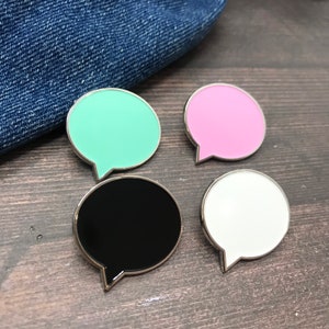 May include: Four enamel pins shaped like speech bubbles. The pins are in black, white, pink, and light green.