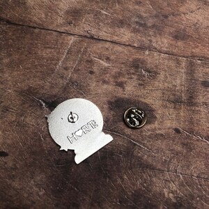 K&ouml;nnte beinhalten: Silberfarbener Metallstift mit einem Globus und dem Text "I &hearts; RT" auf der R&uuml;ckseite. Der Stift hat einen Standard-Schmetterlingsverschluss.