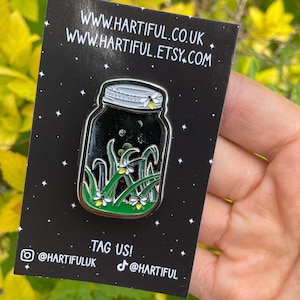 May include: A black enamel pin featuring a jar with fireflies and grass. The jar has a silver lid and a black interior with glowing green fireflies. The pin is on a black card with the website address and social media handles.