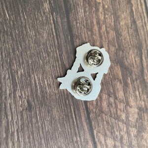 May include: A silver-colored enamel pin with two pin backs. The pin has a floral design. The pin is on a wooden surface.