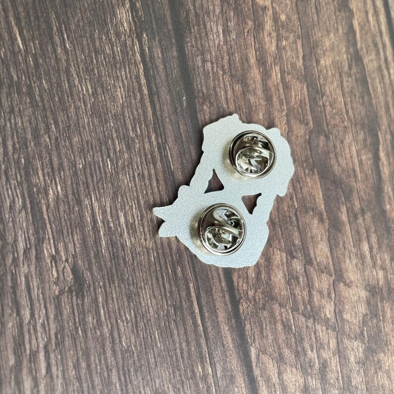 May include: A silver-colored enamel pin with two pin backs. The pin has a floral design. The pin is on a wooden surface.