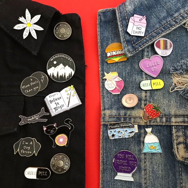 K&ouml;nnte beinhalten: Zwei Jeansjacken mit einer Vielzahl von Emaille-Pins. Die Pins zeigen verschiedene Designs, darunter eine Katze mit einem Donut, ein Buch mit den Worten "Glaube an die Magie", ein Flugzeug, ein Hund, eine Pille, eine Bergkette, ein Feenstaubglas, ein Schild mit der Aufschrift "Keine Milchprodukte", ein Burger, ein Herz mit den Worten "Cheer Up", eine Pille mit den Worten "Chill Pill", ein Fisch in einer T&uuml;te, eine Weltkugel mit den Worten "Du wirst die Welt bereisen" und eine Erdbeere.