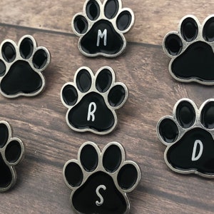 Puede incluir: Seis pines de huella de pata negra con ribete plateado. Cada pin tiene una letra blanca en el centro: M, R, D, S.