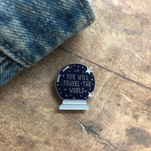 K&ouml;nnte beinhalten: Eine silberne und schwarze Emaille-Anstecknadel in Form einer Kristallkugel mit dem Text "You will travel the world" im Inneren. Die Anstecknadel hat eine wei&szlig;e Basis und zwei silberne Sterne.