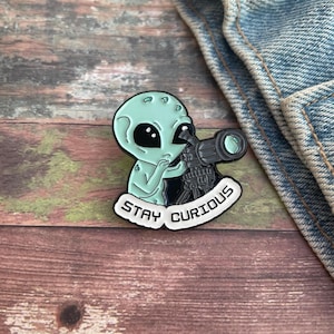 May include: Green alien enamel pin with a telescope and the text "Stay Curious".