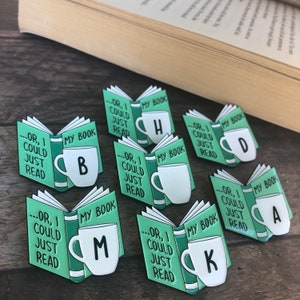 May include: Set of eight enamel pins featuring a green book with a white mug. Each mug has a different letter on it, spelling out the word "BOOK". The text on the book reads "...or, I could just read my book".