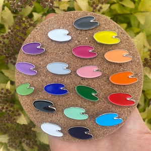 May include: A corkboard with 18 small enamel pins shaped like paint palettes. The pins are in various colours, including red, orange, yellow, green, blue, purple, pink, white, black, and grey.